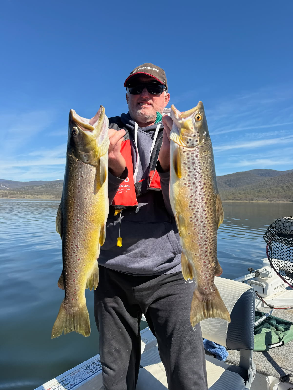 VFC member on a boat holding two large brown trout with a lake and mountains behind